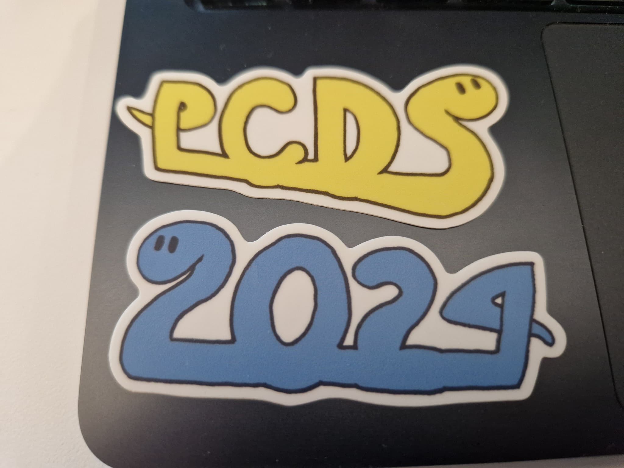 Two stickers. One yellow snake with its body looping and spelling out the letters PCDS (for Python Core Dev Sprint), one blue snake spelling 2024.