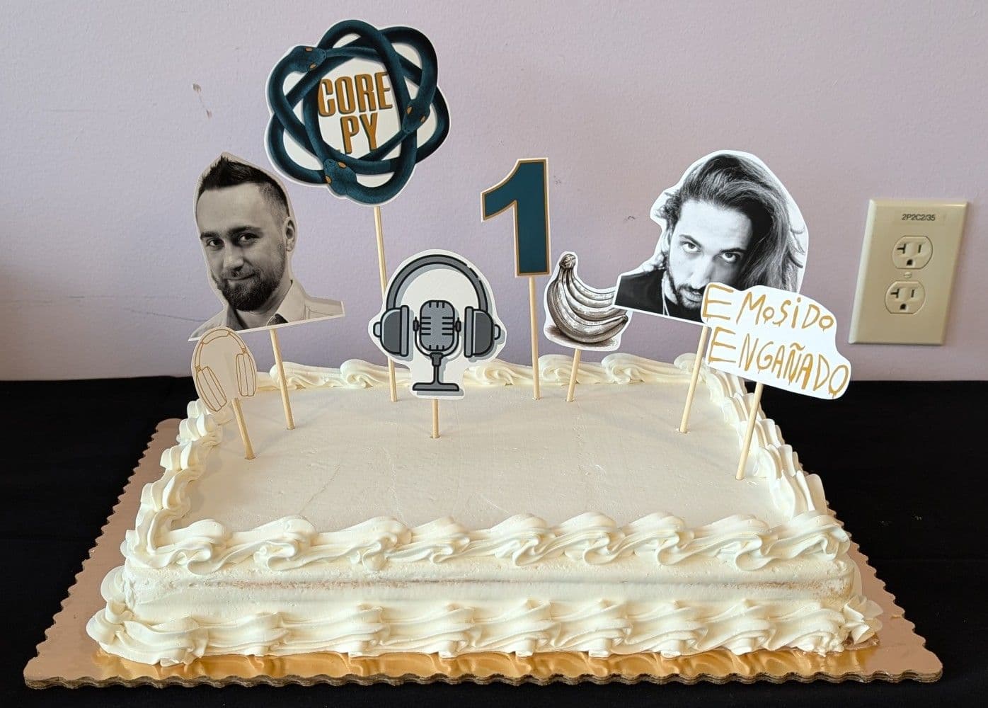 A big white cake, with decorations of Łukasz and Pablo's faces, along with the Core.py logo, a big digit 1, headphones, microphone, bananas, and "emosido engañado" graffiti.