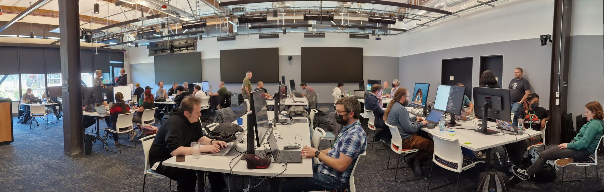 A panoramic shot of a room of people, some sitting at desks, some standing, some working on laptops, some in discussion.